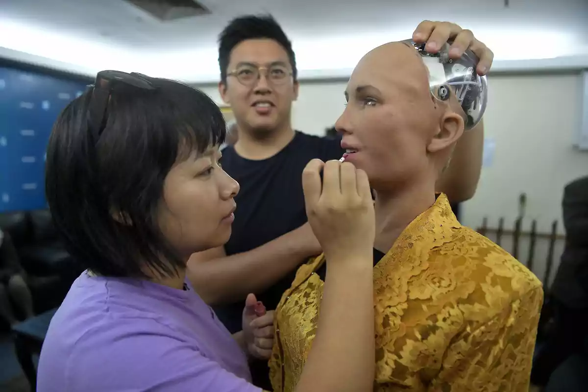 Dues persones ajusten i maquillen un robot humanoide en una habitació.