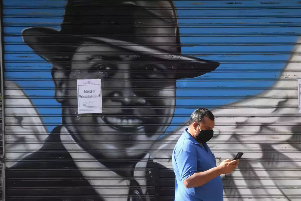 Un home amb màscara i camisa blava revisa el telèfon davant d'una porta metàl·lica amb un mural d'un home amb barret i ales, i un cartell que diu "Estem a Valentín Gómez 2643".