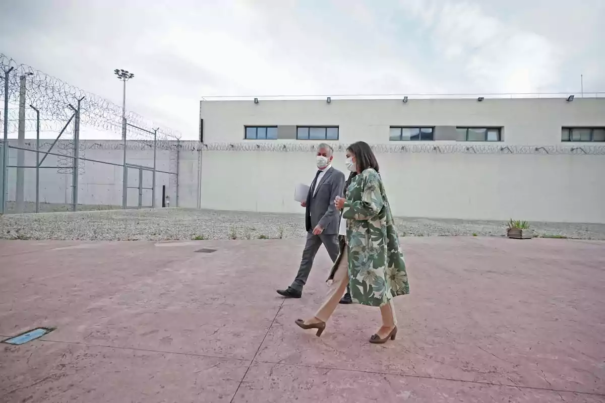 Dues persones amb màscares caminant al pati d'una presó amb filferro de pues i un edifici de fons.