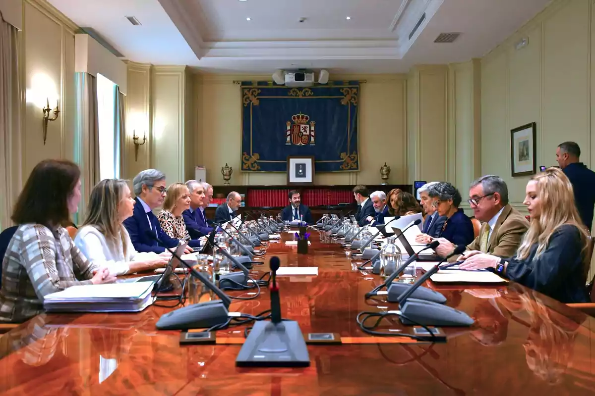 Persones assegudes al voltant d'una taula de conferències en una sala de reunions formal, amb micròfons i documents davant seu, i un escut d'armes a la paret del fons.