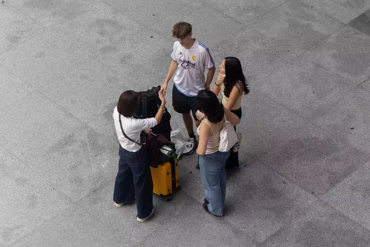 Un grup de quatre persones, tres dones i un home, estan drets en un espai obert de terra grisa, amb una maleta groga i diverses bosses al seu voltant.