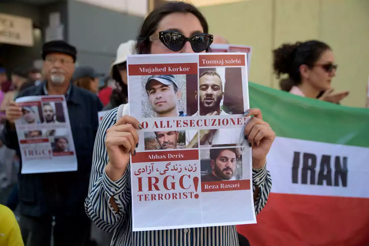 Una persona sosté un cartell amb fotos i noms de diverses persones, amb un missatge en italià que diu "NO ALL'ESECUZIONE!" i un altre missatge en anglès que diu "IRGC TERRORIST!" mentre participa en una protesta; al fons, altres persones també sostenen cartells i una bandera de l'Iran.