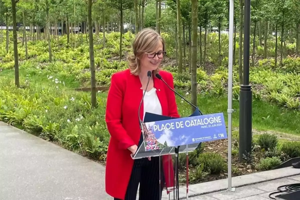 Una dona amb ulleres i jaqueta vermella està parlant en un podi a l'aire lliure en un parc, amb un cartell que diu Place de Catalogne i la data 23 juin 2024.
