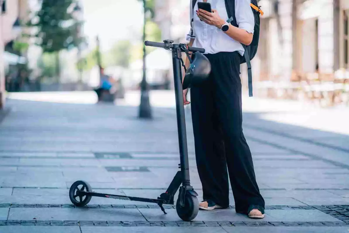 Persona de peu al carrer amb un patinet elèctric i usant un telèfon mòbil.