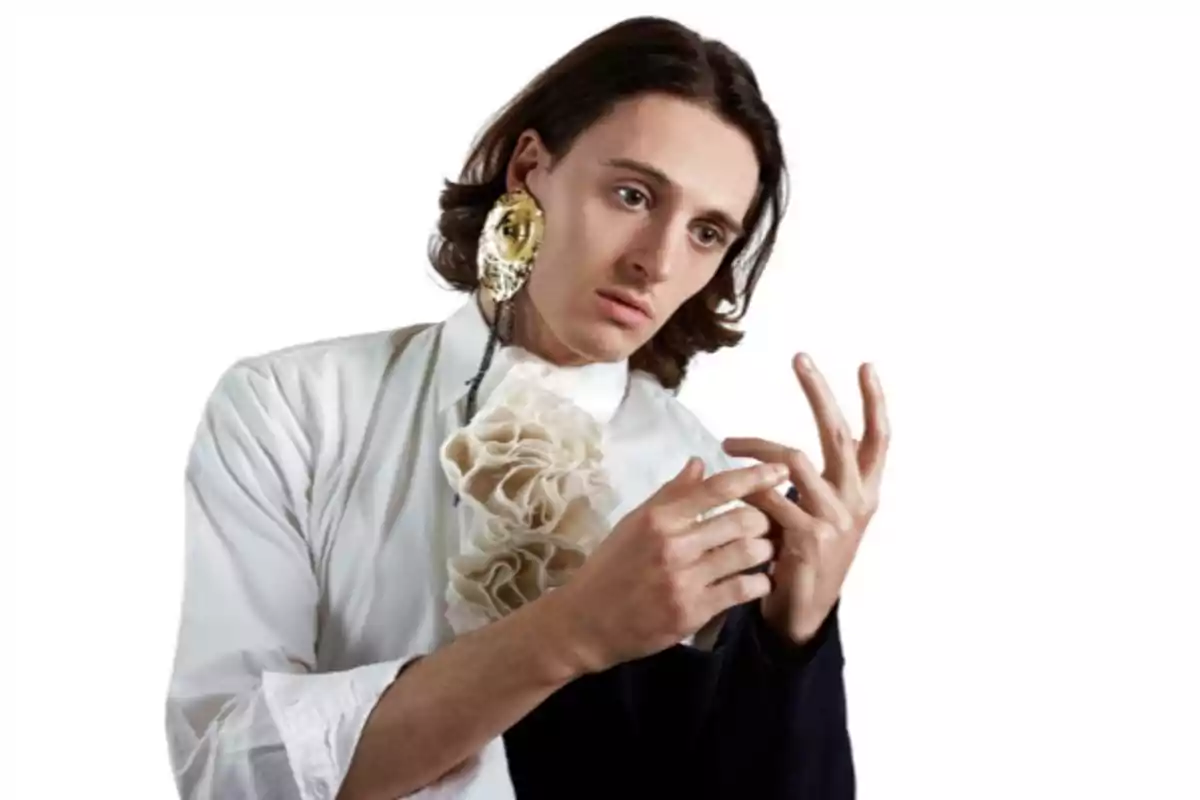 Un home amb cabell llarg i fosc, vestit amb una camisa blanca amb un adorn de volants al pit, porta un pendent gran i daurat a l'orella esquerra. Està mirant les mans amb una expressió pensativa. El fons és blanc.