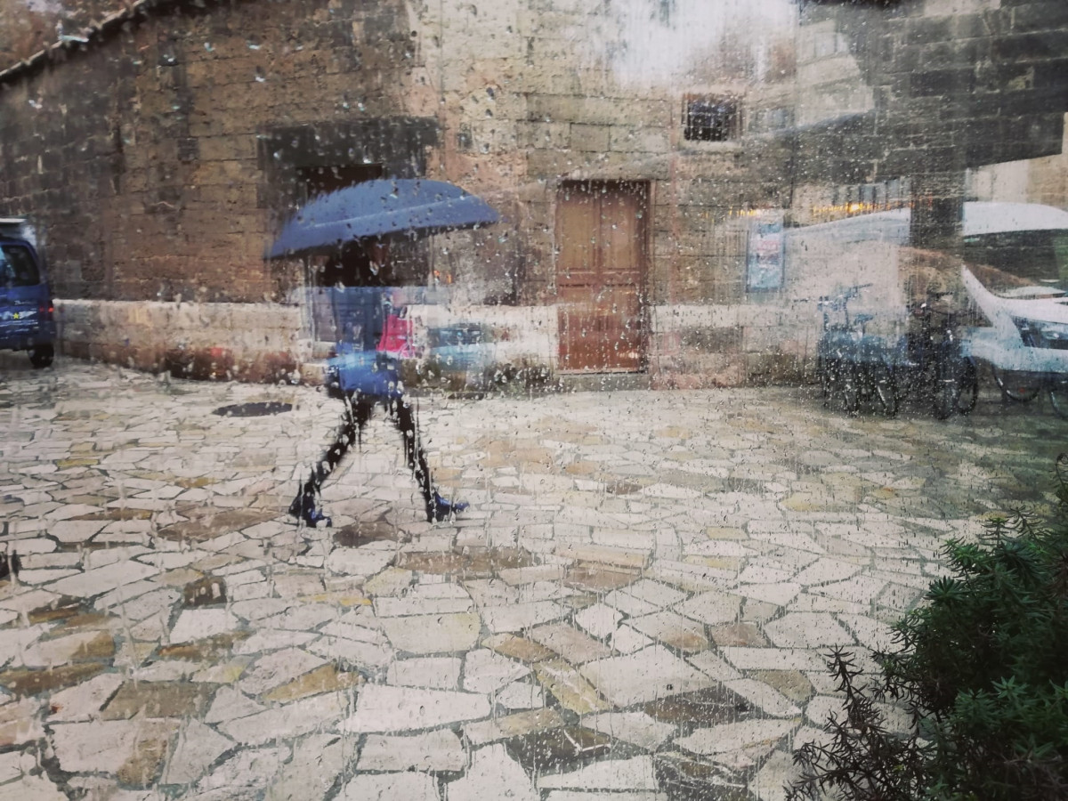 Una dona passeja pel centre de Palma amb un paraigua, vista a través d'un vidre amb gotes de pluja.